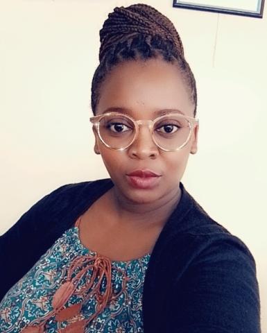alt=" A woman with braided hair in a bun, wearing glasses and a patterned top with a black cardigan, looking at the camera with a neutral expression